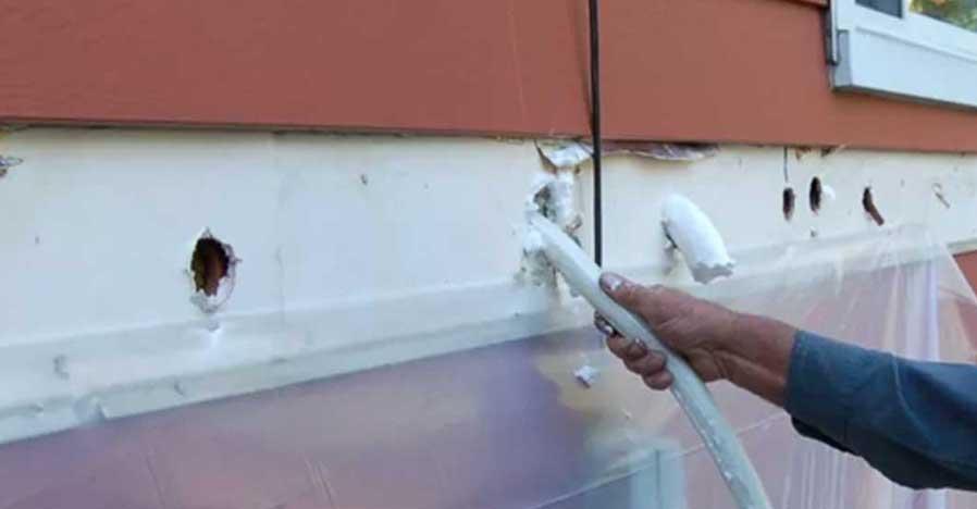 Person insulating a wall by injecting foam through small holes at a building site.